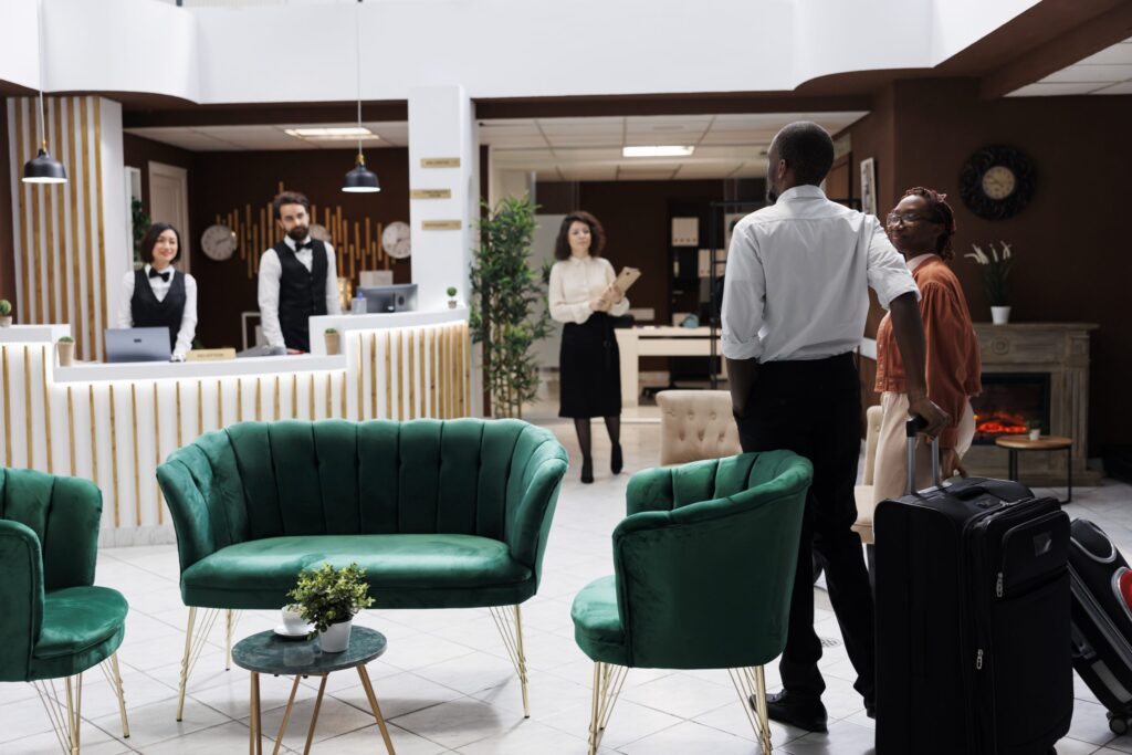 A wide shot of a hotel lobby with a couple standing at the front desk with luggage. Two hotel staff members are behind the counter, and another employee is standing in the background with a tablet. There are two green velvet armchairs and a small table in the foreground.