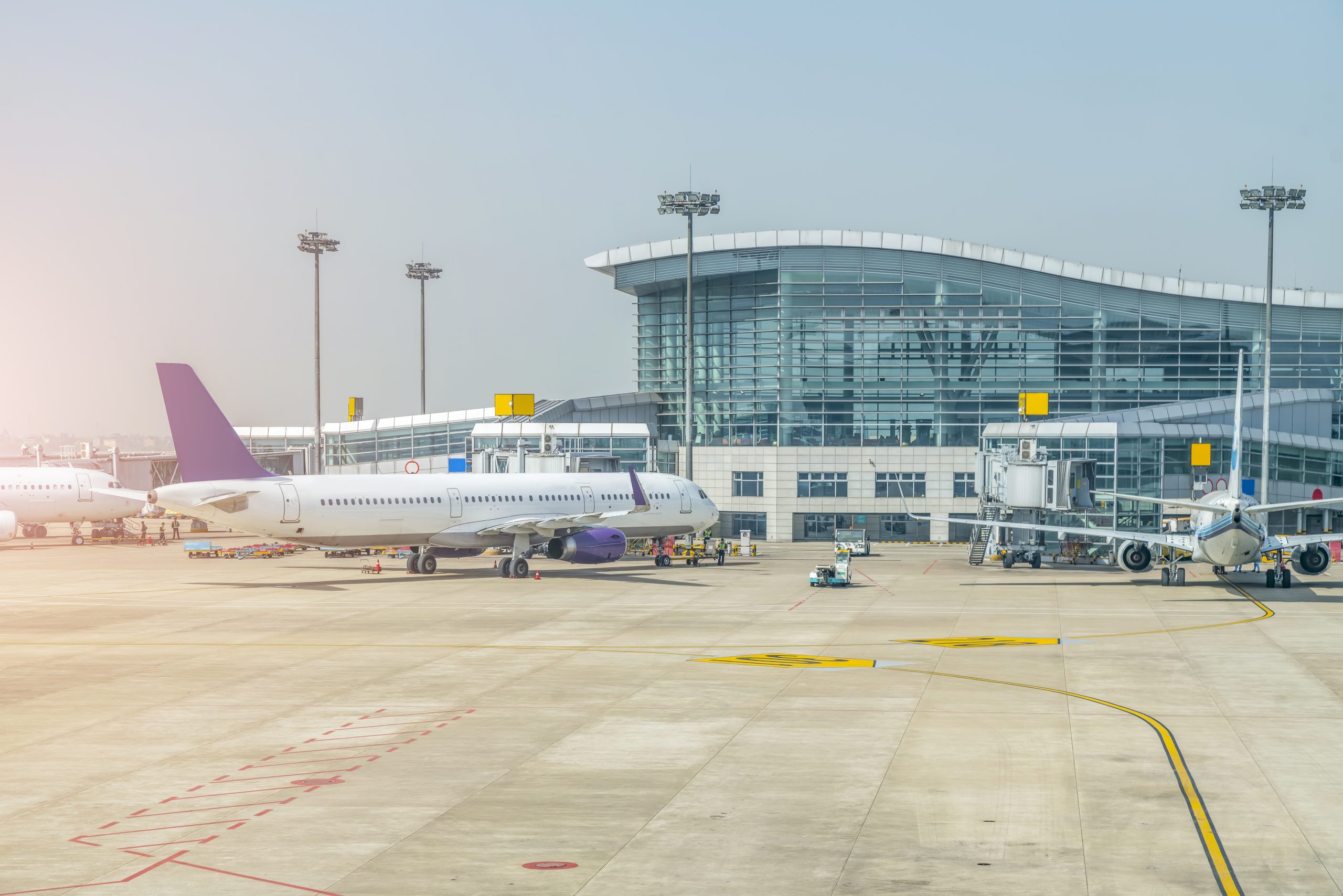 A view of a busy airport tarmac on a bright day. Several airplanes are parked near a large, modern terminal building with a glass façade and curved roof.