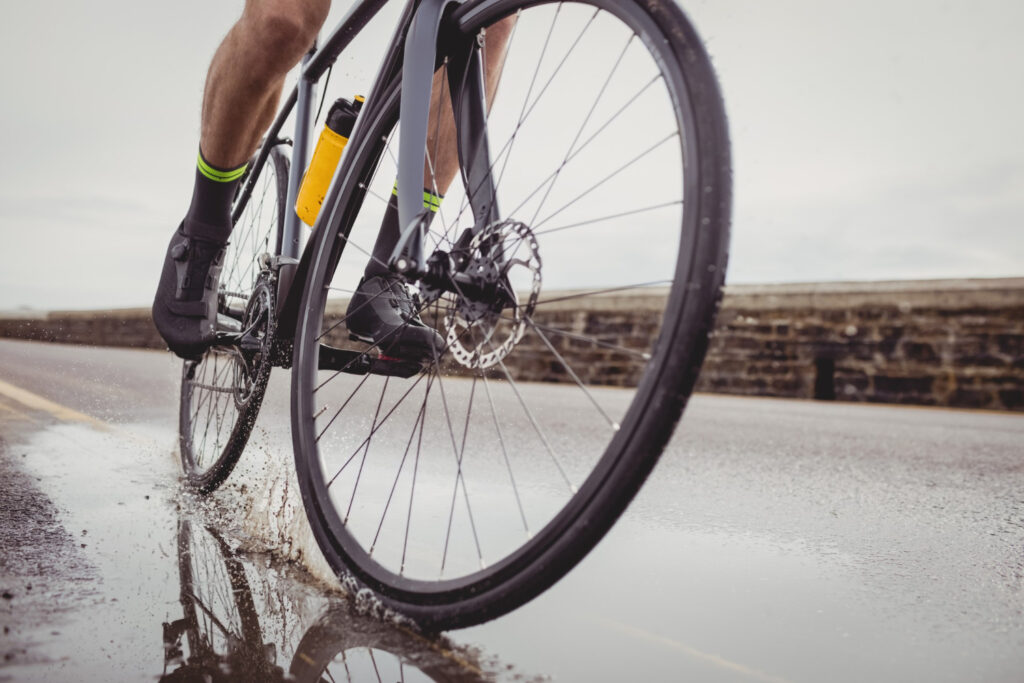 A low-angle close-up of a person on a bicycle riding through a puddle on a wet road. The wheels splash water as they move, and a water bottle is in the bike frame.