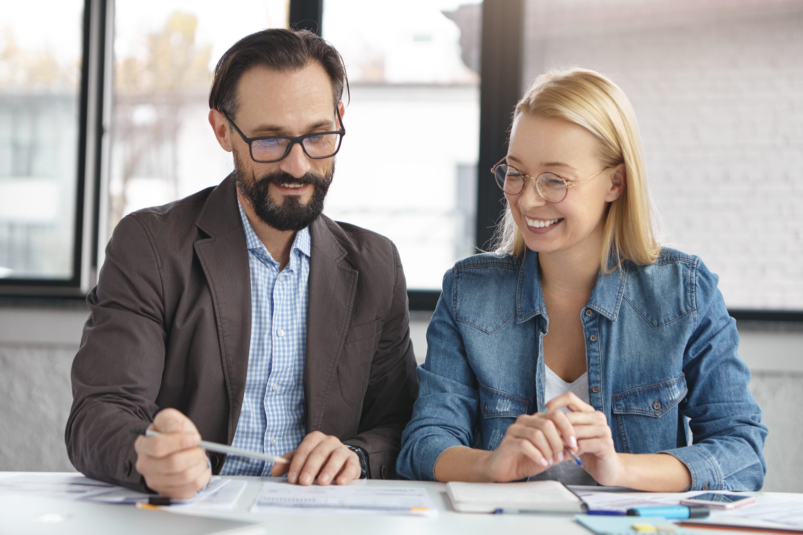 A man and a woman wearing glasses are sitting at a table with papers in front of them, looking down and smiling. The woman is wearing a denim jacket and the man is wearing a brown blazer.