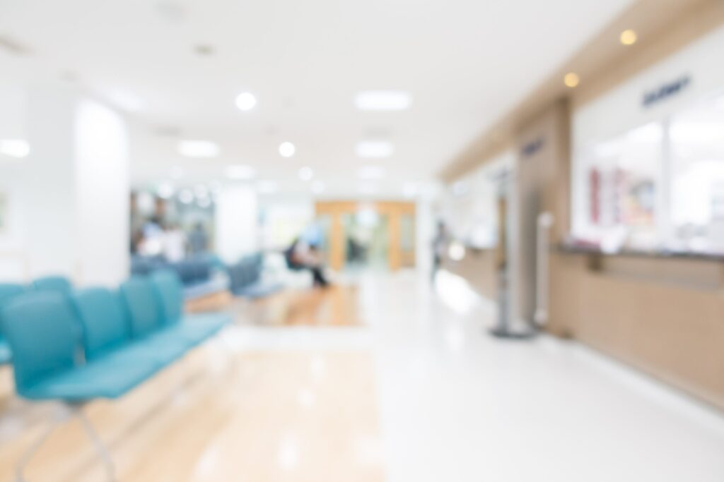 A deliberately blurred or out-of-focus image of a bright, clean hospital or clinic waiting area with a row of turquoise chairs on the left and a reception counter in the distance.