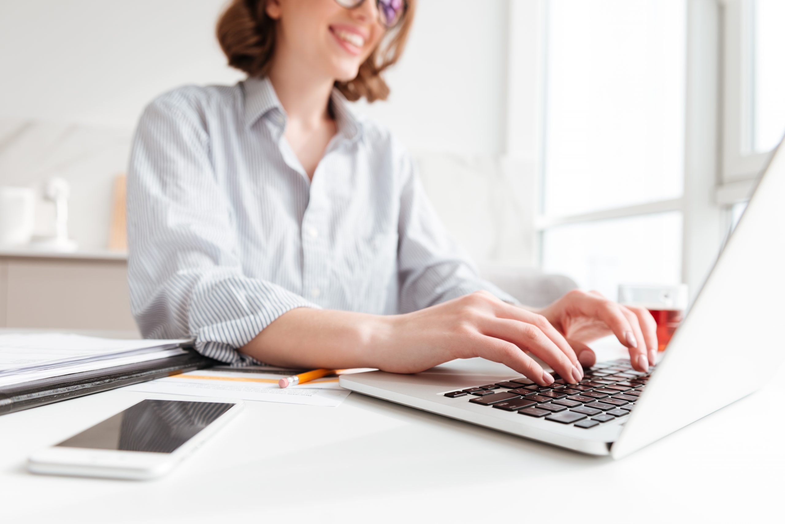 A smiling woman wearing glasses types on a laptop at a desk.