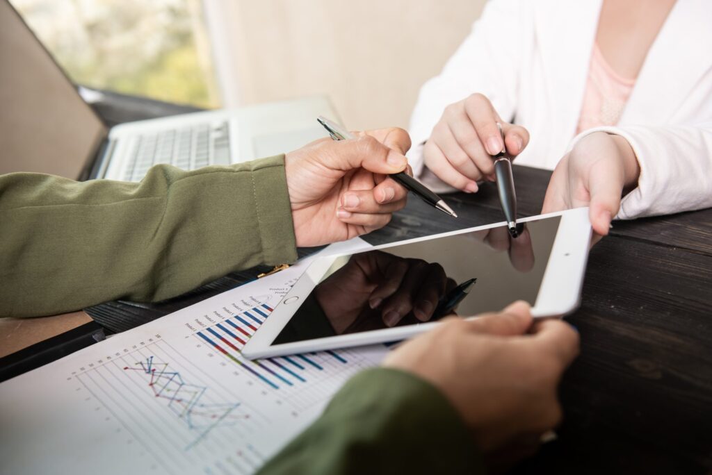 Two people are in a meeting, with one holding a tablet and another pointing to the screen with a pen. Papers with charts and graphs are on the table, and a laptop is visible in the background.