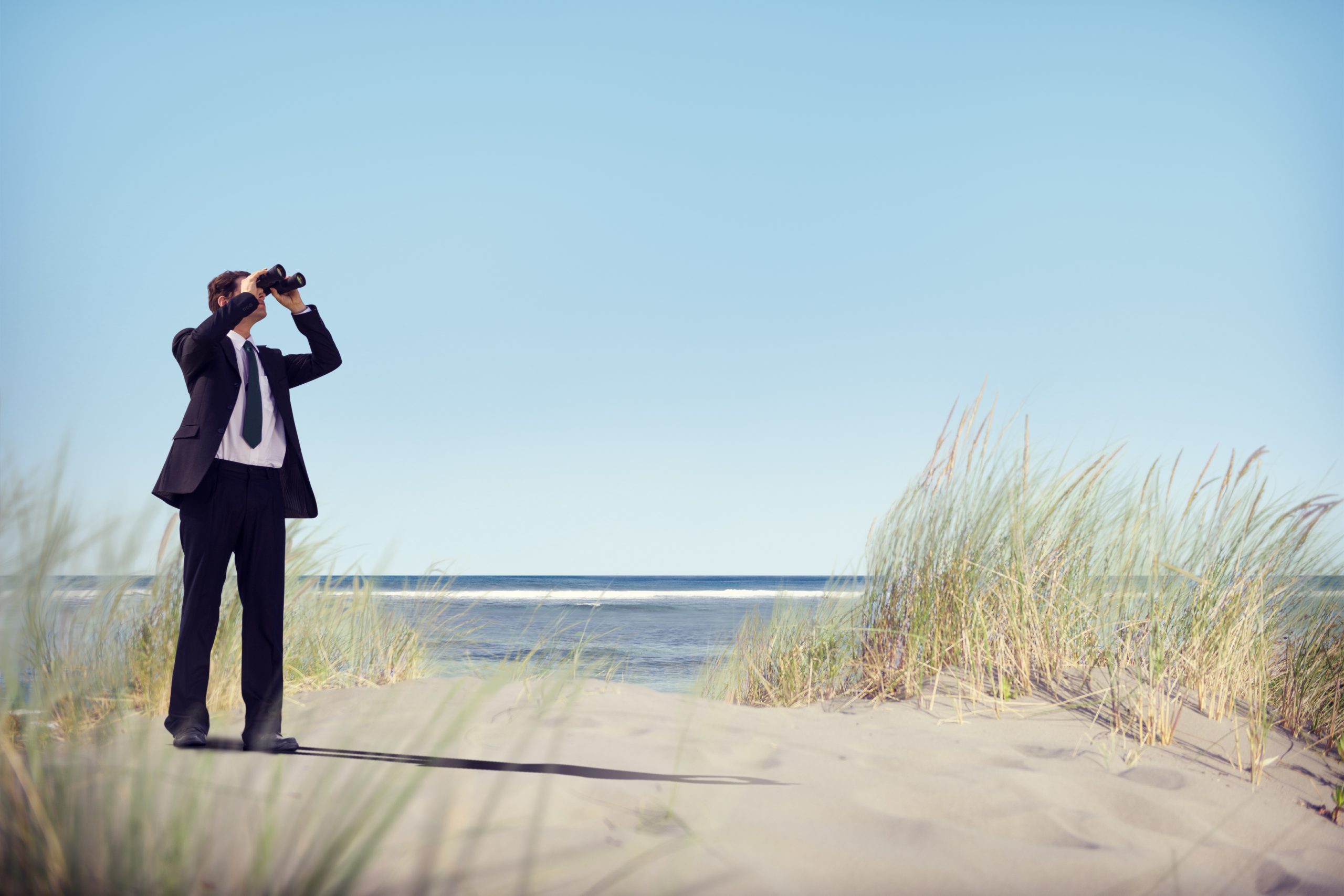 A businessman stood on the sand at the beach with binoculars looking to the sky.