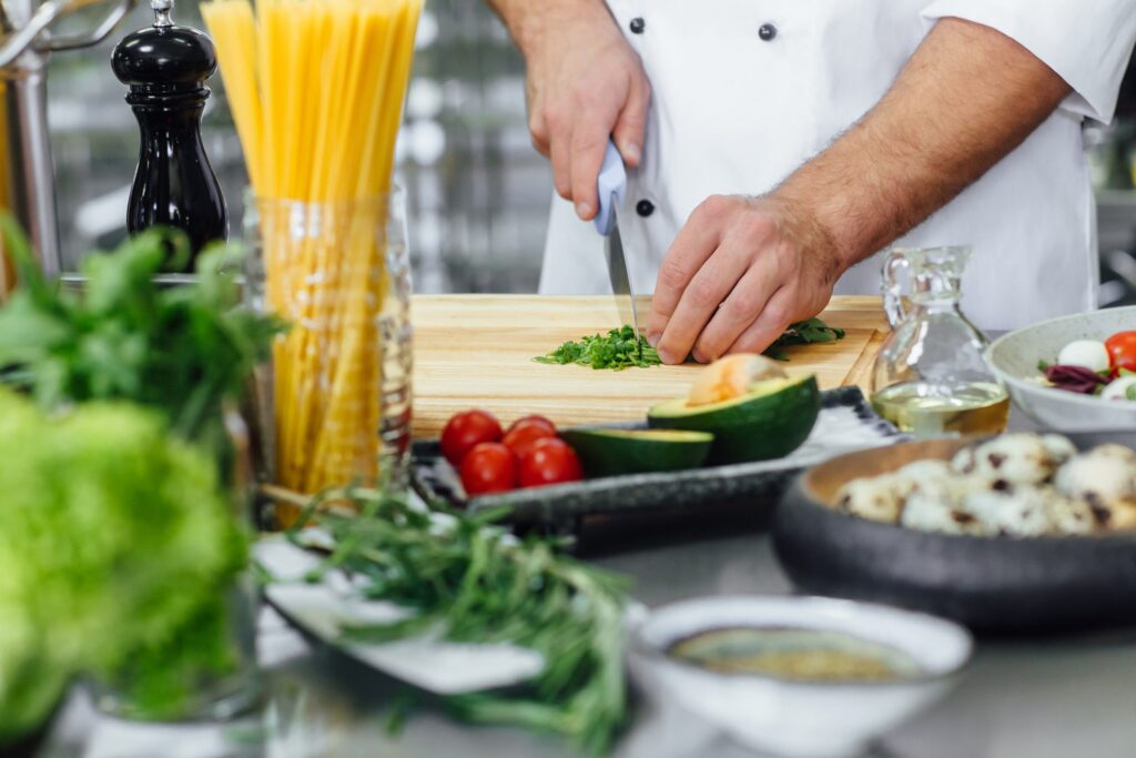 A close-up of a chef chopping fresh herbs on a wooden cutting board in a professional kitchen. Various ingredients, including uncooked pasta, cherry tomatoes, and quail eggs, are on the counter.