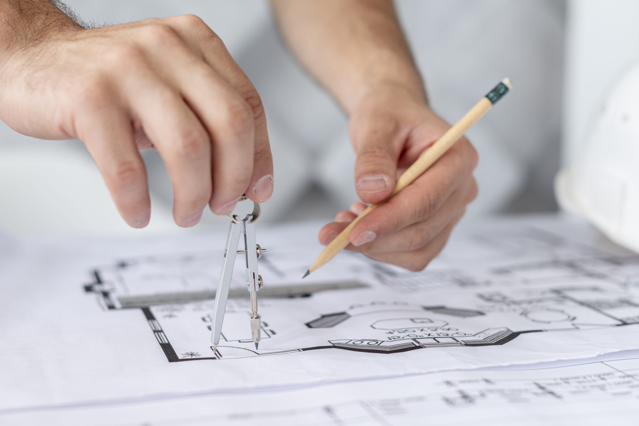 A person's hands are using a compass and a pencil to draw on a blueprint or architectural plan. A white hard hat is partially visible in the background.