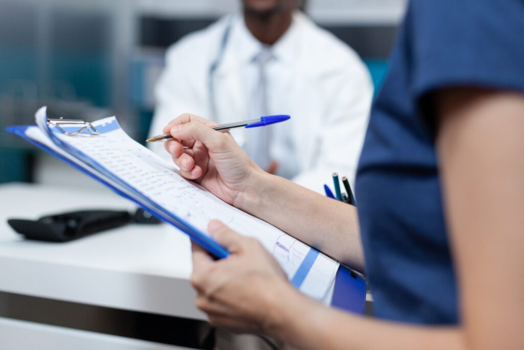 A nurse in blue scrubs holds a clipboard and writes with a blue pen. In the blurry background, a doctor in a white coat sits at a desk.