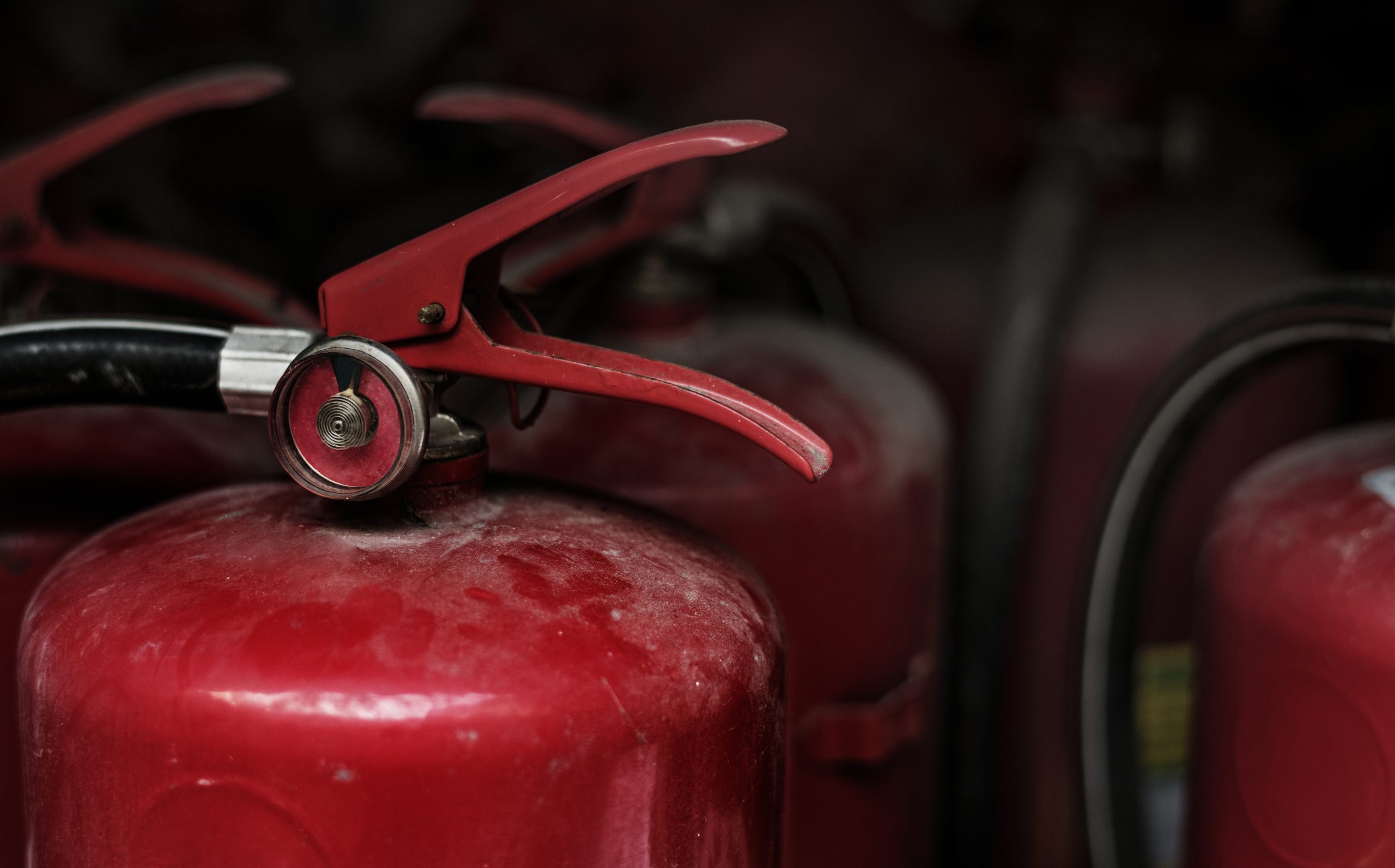 A close-up, high-angle shot of several red fire extinguishers lined up. The focus is on the top of the nearest extinguisher, showing the red handle, black hose, and pressure gauge.