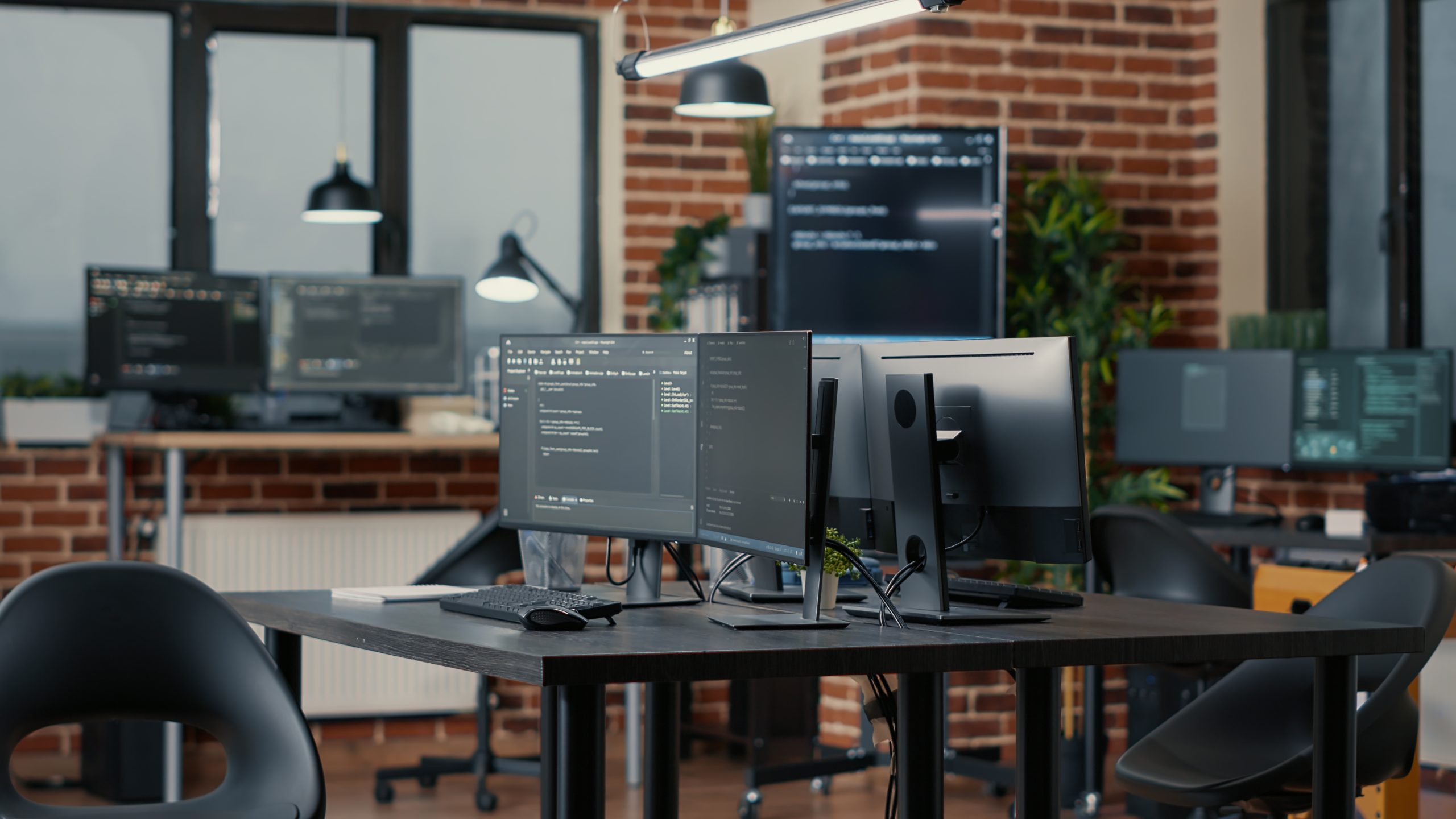 An empty office with two computer monitors displaying lines of code. The room has a red brick wall and multiple desks with more computers in the background.