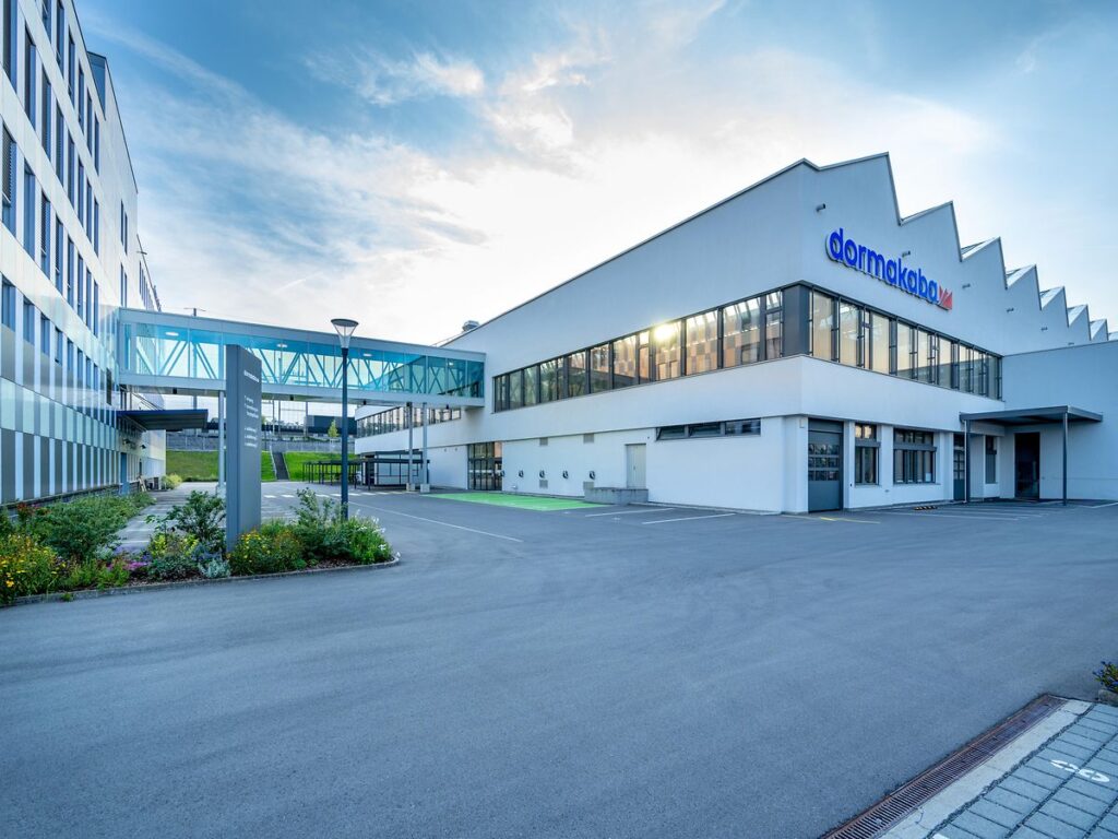 A modern, white corporate building with a skywalk connecting to another building, with the "dormakaba" logo visible on the side.