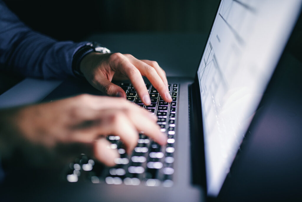 A person's hands are typing on a glowing laptop keyboard in a dark room.