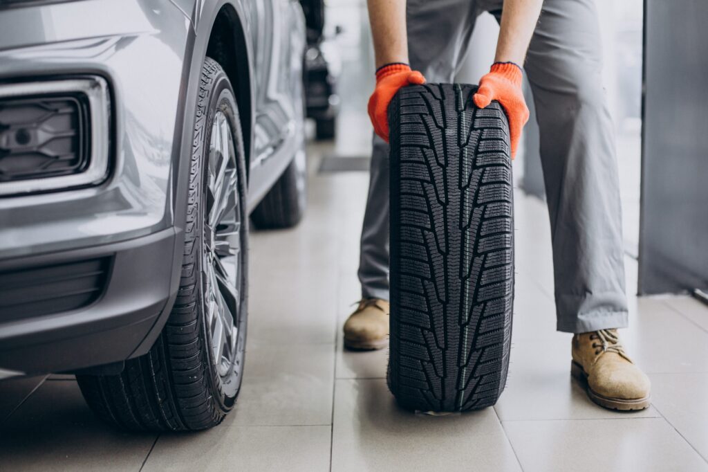 A mechanic wearing orange gloves holds a new car tire in a garage.