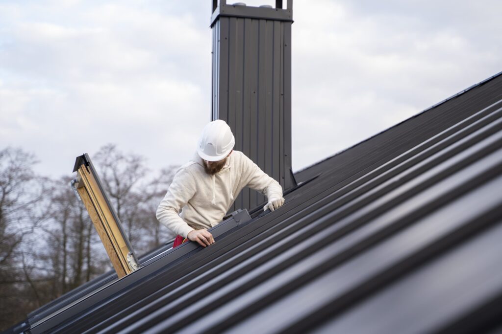 A man with a white hard hat and beard is working on a black metal roof. A chimney is also visible on the roof and trees are in the background.