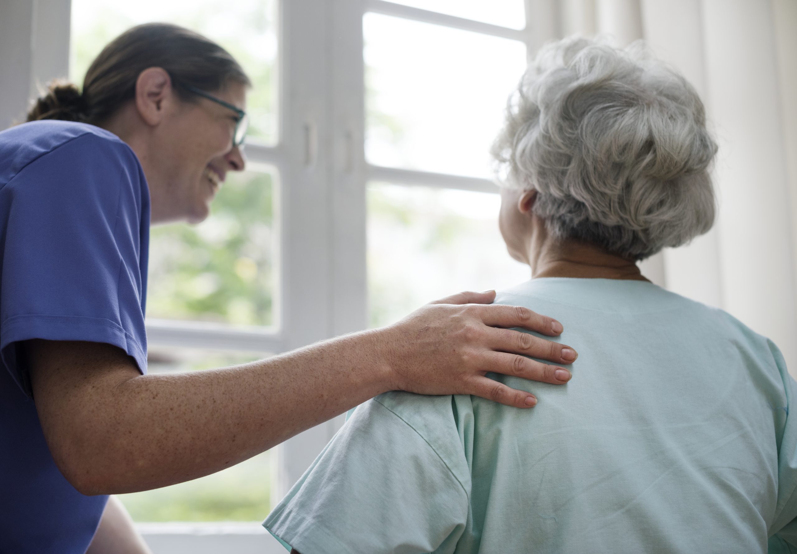 A nurse in a blue uniform stands and smiles at an elderly patient, placing her hand gently on the patient's shoulder. The patient, who has grey hair, is seen from the back, sitting and looking out a large window.