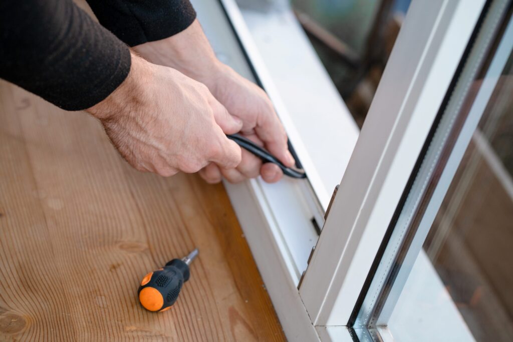 A person's hands install a black rubber gasket onto a white window frame, with a screwdriver nearby.