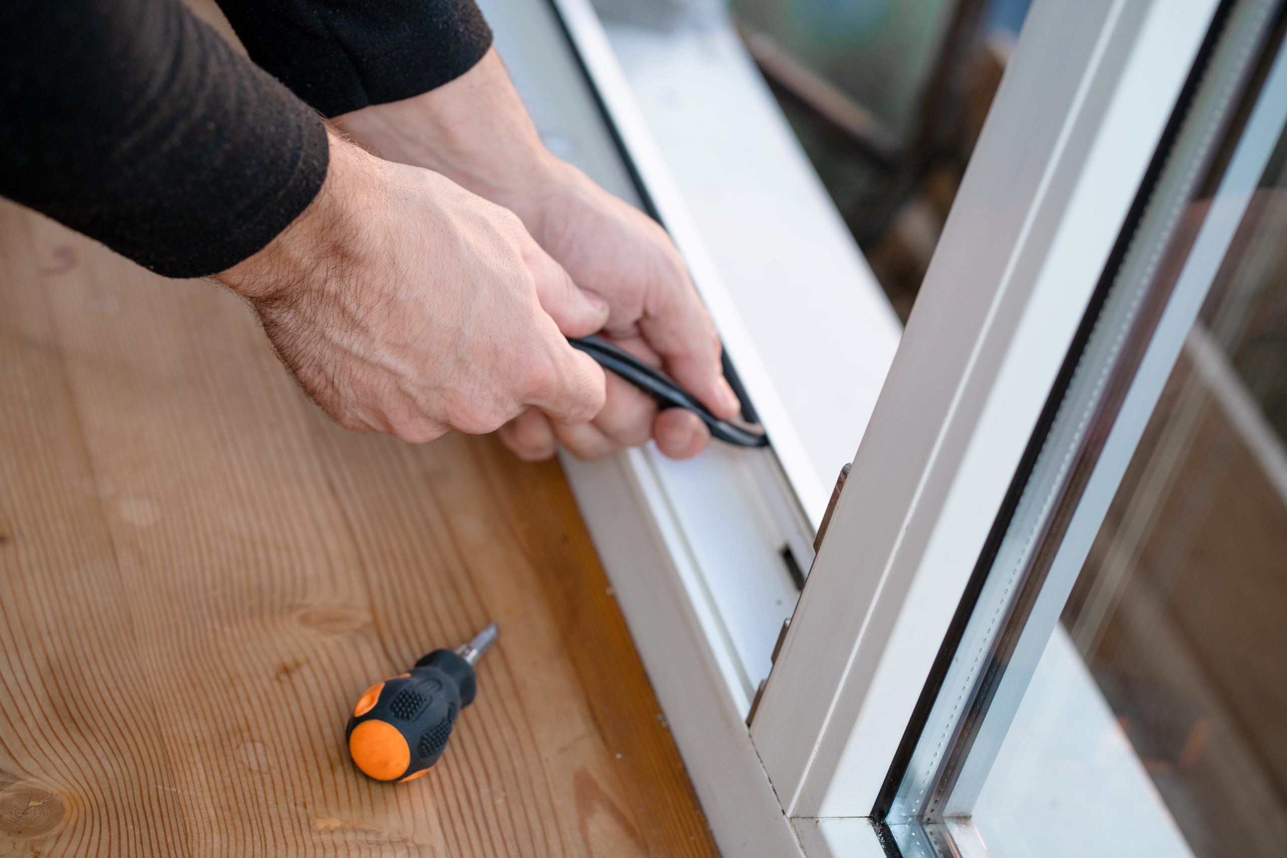 A person's hands install a black rubber gasket onto a white window frame, with a screwdriver nearby.