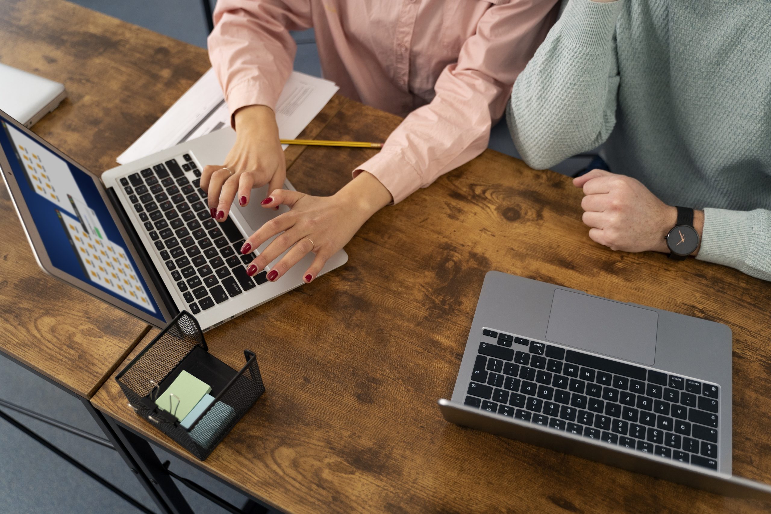 An overhead shot of two people working at a wooden desk with two laptops. One person's hands, with red nail polish, are typing on a laptop, while the other person's hands are resting on the desk.