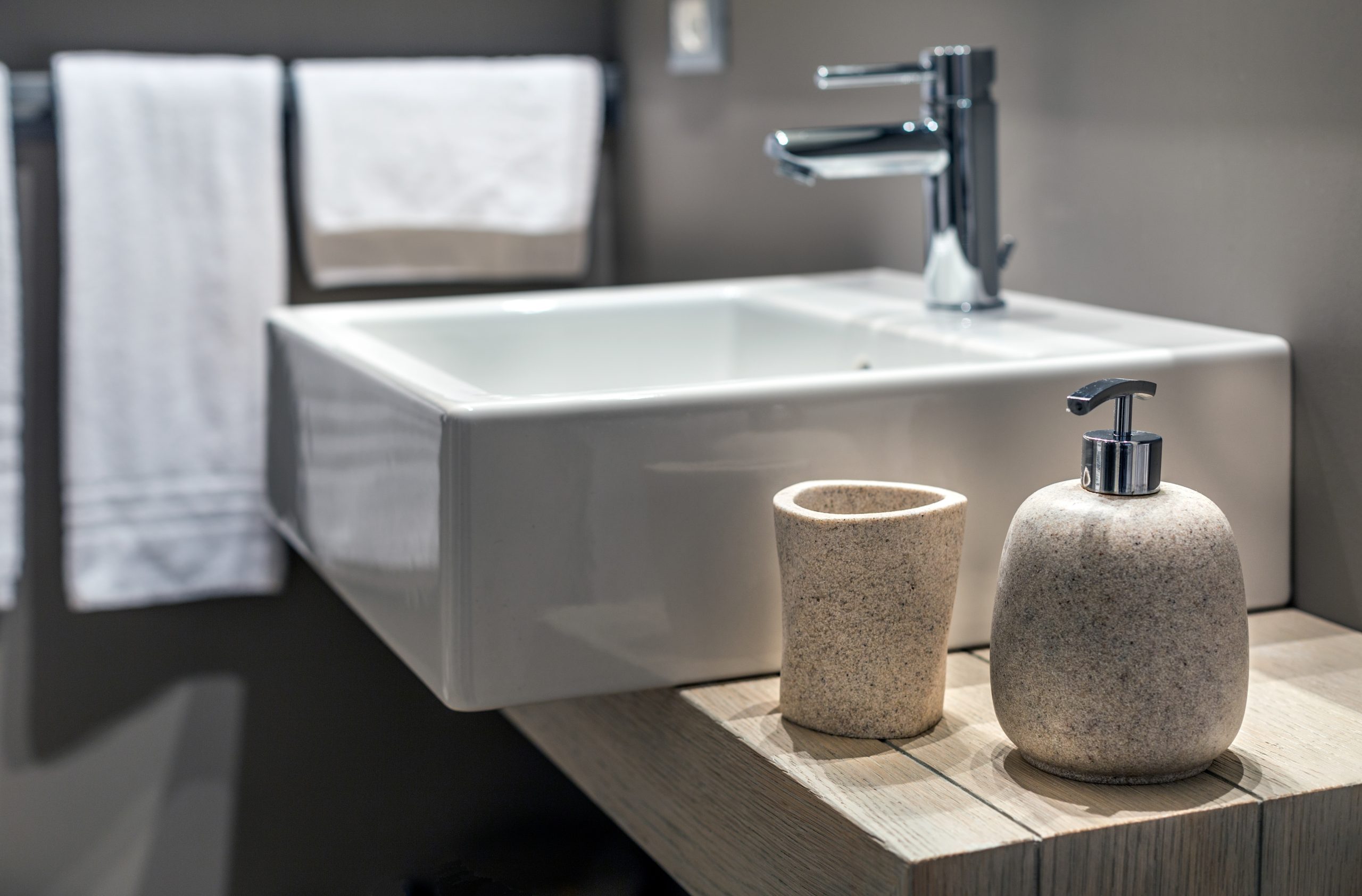 A clean, modern bathroom with a white rectangular sink. A tap with a single handle is attached to the wall, and two white towels hang on a rack in the background. In the foreground, a speckled beige soap dispenser and toothbrush holder sit on a wooden countertop.