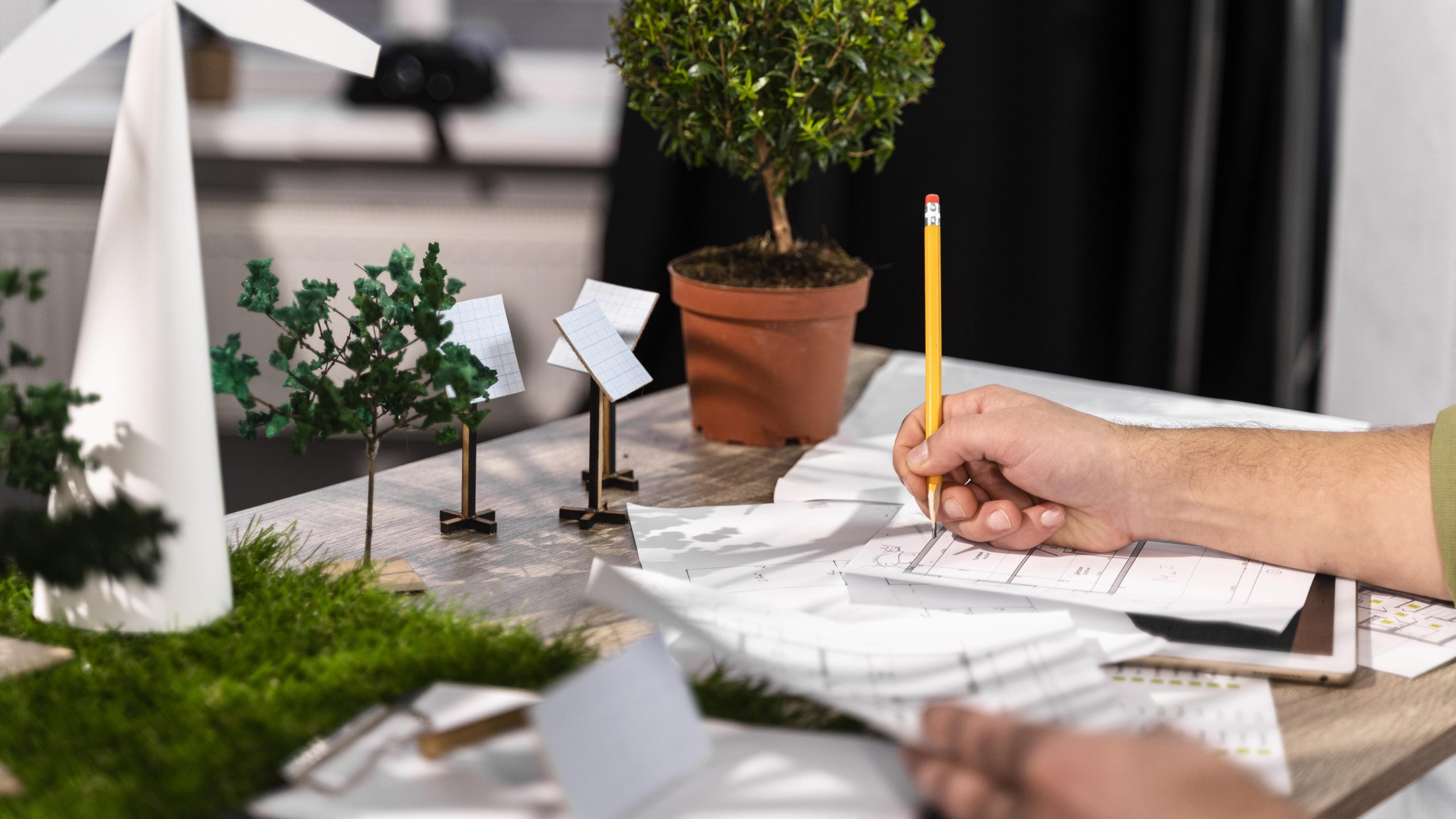 A man's hand holds a pencil over a blueprint on a desk, next to a variety of small models, including a white wind turbine, green trees, and solar panels on tiny poles. A small potted plant and other documents are also on the desk.