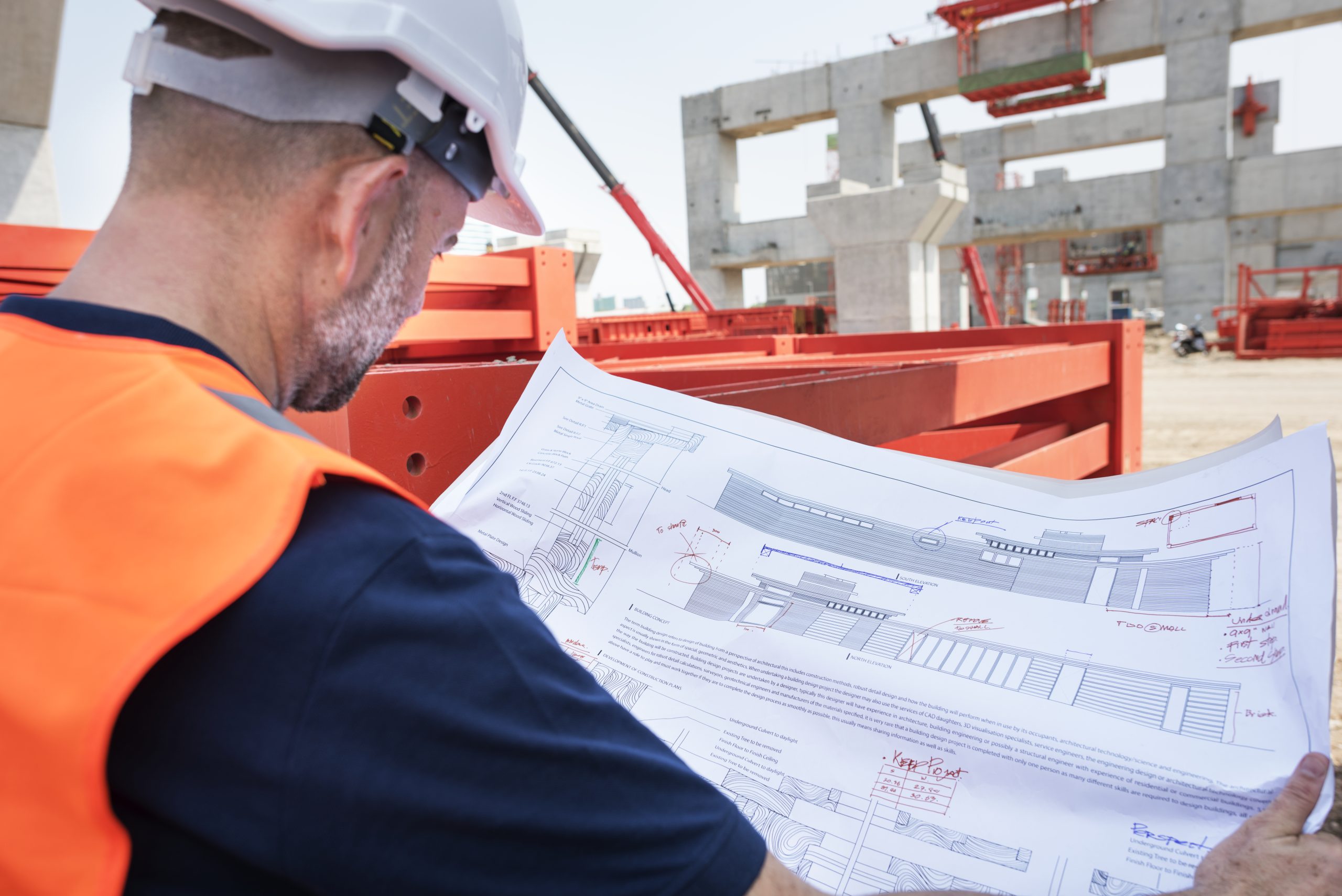 A construction worker wearing a hard hat and safety vest is looking at a set of blueprints at a construction site with concrete structures and a crane in the background.