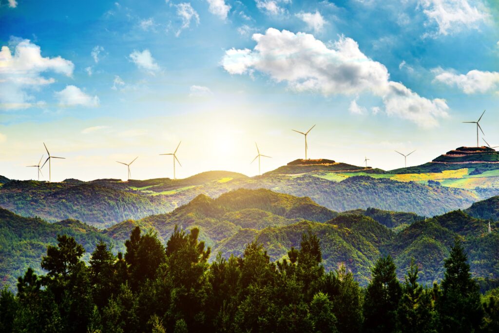 A landscape shot of a lush green mountain range with a few scattered wind turbines on the peaks, silhouetted against a bright sun and a vibrant blue sky with scattered clouds. The foreground shows dense, dark green treetops, while the background reveals rolling hills and the sun's rays peeking through the clouds.