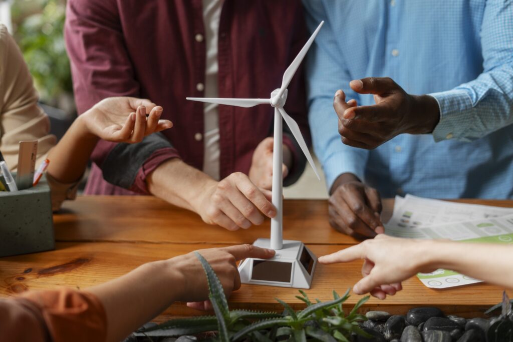 A medium shot of four people's hands and arms surrounding a small, white model of a wind turbine on a wooden table. Two hands point toward the base of the model, which has solar panels on the sides, while another person's hand reaches out as if to touch it. In the foreground, there are small plants and rocks on the table.