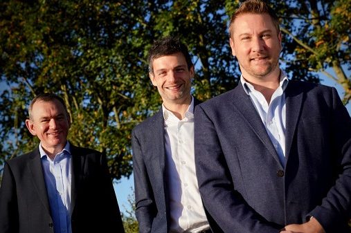 Three men in business casual attire smile while standing outdoors.