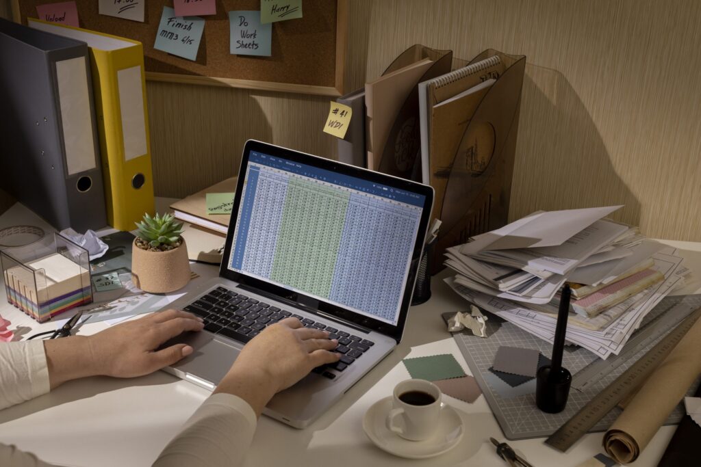 A person's hands are typing on a laptop with a spreadsheet on the screen. The cluttered desk also holds folders, sticky notes on a corkboard, a stack of papers, a coffee cup, and various office supplies.