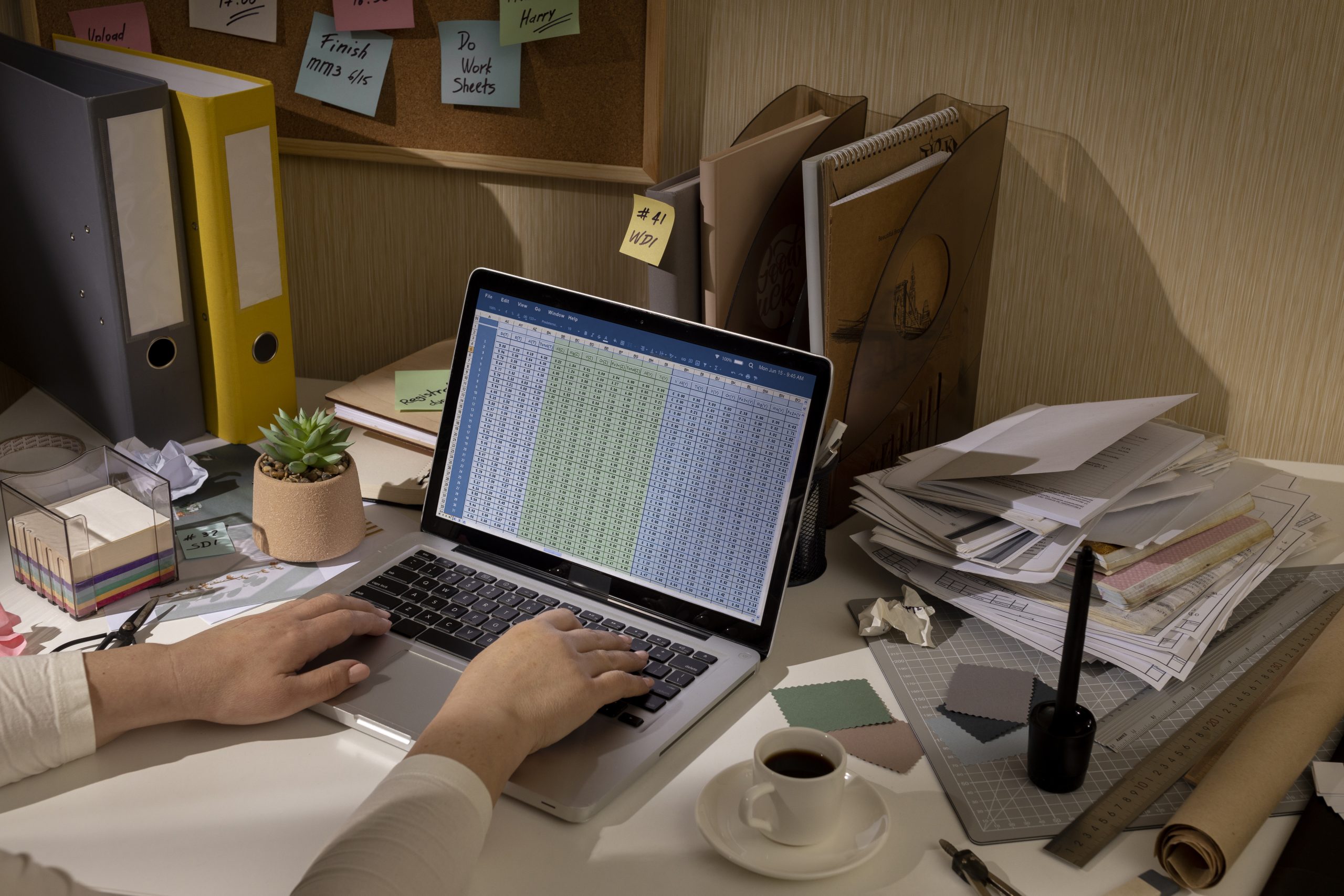 A person's hands are typing on a laptop with a spreadsheet on the screen. The cluttered desk also holds folders, sticky notes on a corkboard, a stack of papers, a coffee cup, and various office supplies.