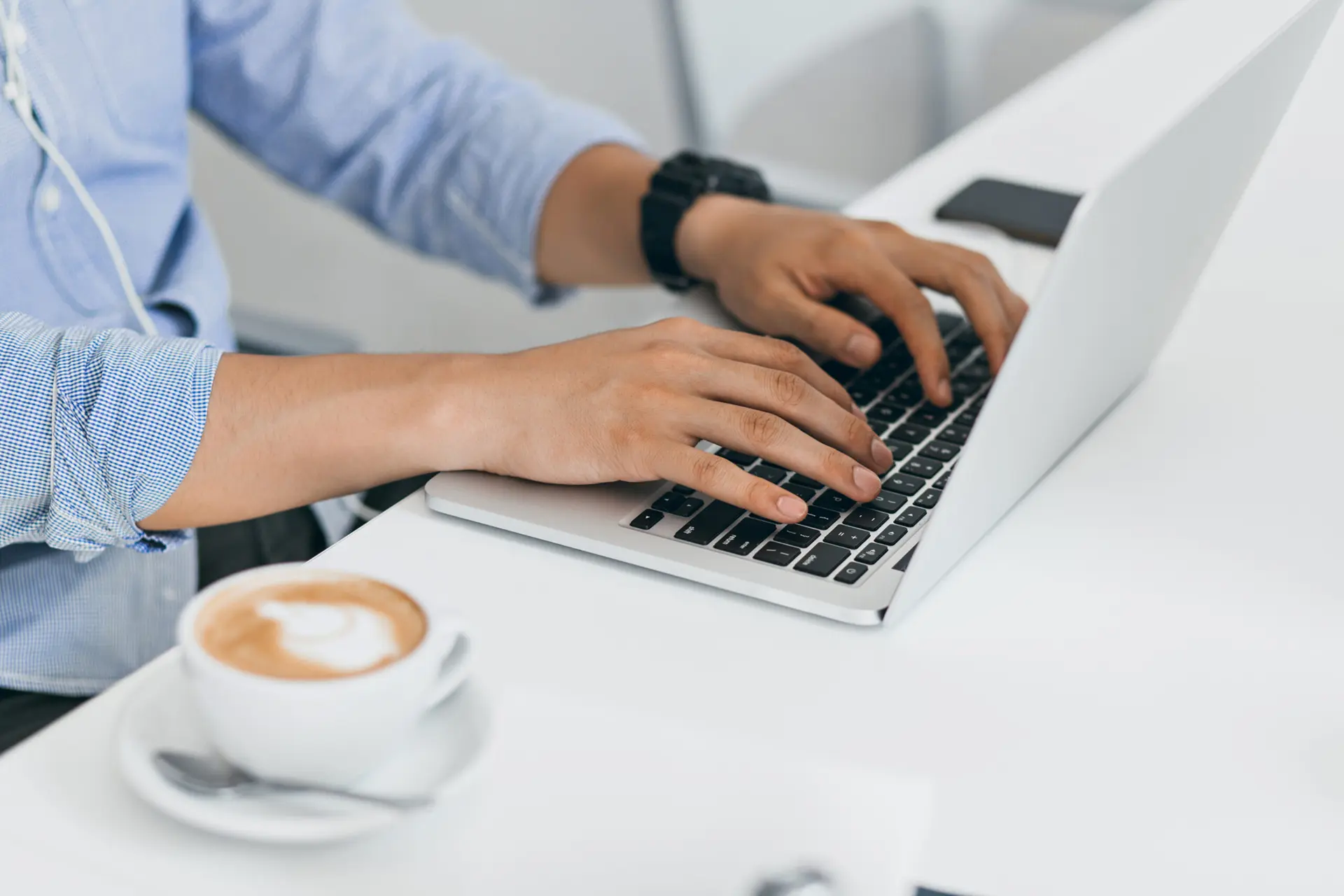 A person in a blue shirt typing on a laptop at a white desk, with a cup of coffee on a saucer nearby in an office setting.